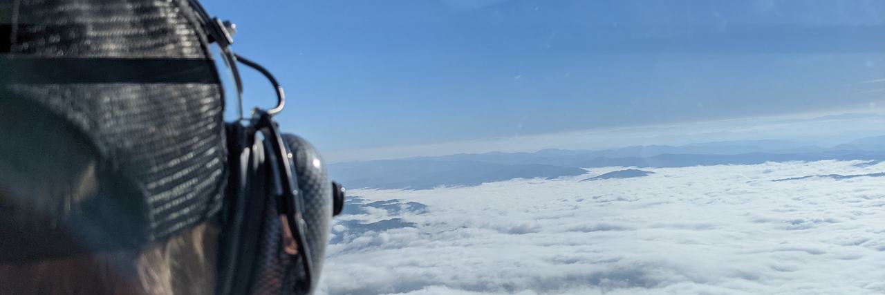 Pohled z kokpitu vírníku: pilot v helmě, pod ním moře mraků a na obzoru hory.